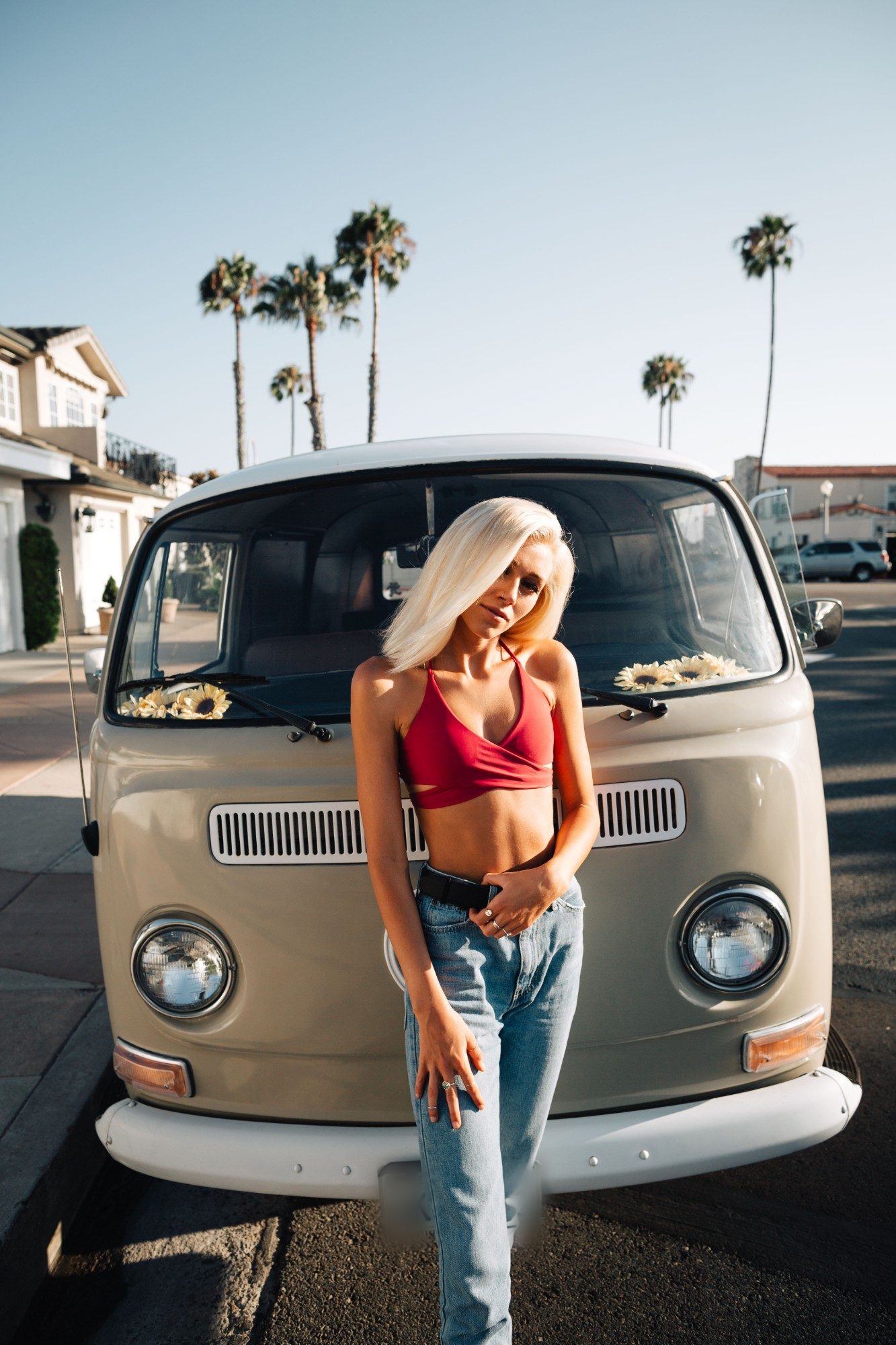 Model in a red top and jeans leaning on the front of a vintage VW bus under palm trees